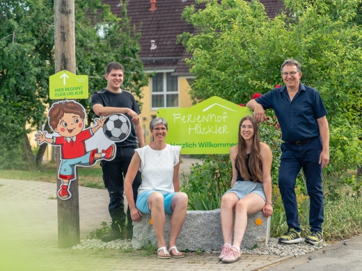 Familie Häckler sitzt und steht am Willkommensschild des Ferienhofs, daneben die Fußballjungen-Figur