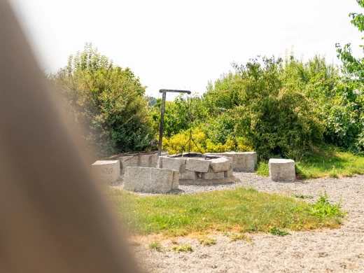 Grillplatz mit Feuerstelle und Sitzblöcken, umgeben von Sträuchern und Wiese
