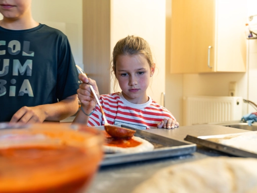 Kind am Tisch beim Pizza backen, vorn eine Schüssel Tomatensauce, links ein Erwachsener im Hintergrund
