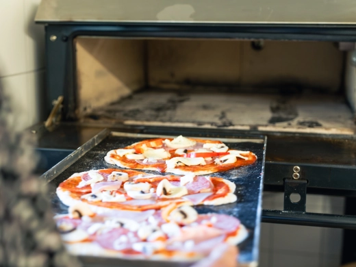Belegte Pizzen auf einem Blech vor der Öffnung des Steinbackofens, bereit zum Backen