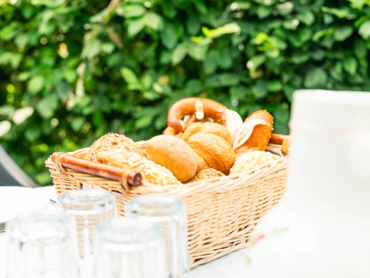 Brötchenkorb und Gläser auf einem weißen Tisch vor einer grünen Hecke
