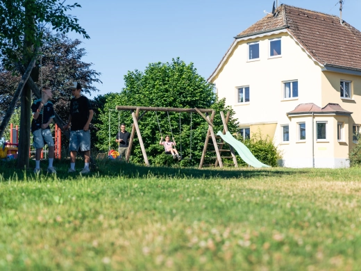 Kind auf der Schaukel mit Rutsche auf dem Gelände des Ferienhofs, davor Wiese, rechts ein gelbes Gebäude