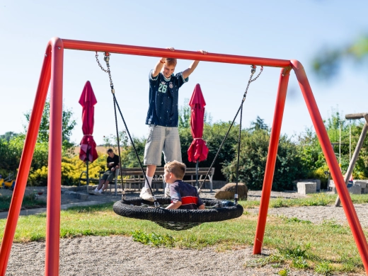 Zwei Kinder an der runden Nestschaukel auf dem Spielplatz, ein Junge steht am roten Schaukelgerüst