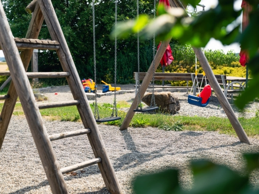 Spielplatz mit Holzgerüst und Schaukeln auf Kiesboden, im Hintergrund weitere Spielgeräte