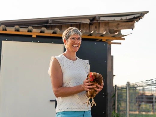 Christine Häckler hält lächelnd ein Huhn im Arm, hinter ihr die Wand des Hühnerstalls