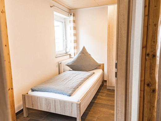 Ferienwohnung 10: Kleines Schlafzimmer mit Einzelbett, Kissen und Fenster, Blick durch die offene Tür