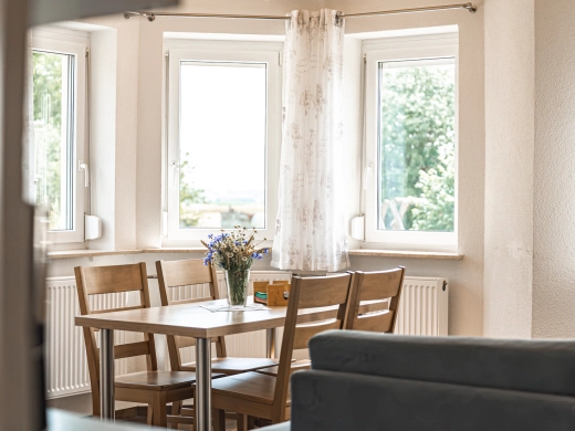 Ferienwohnung 10: Esstisch mit Holzstühlen vor dem Erkerfenster, Sofa im Vordergrund, Blick ins Grüne