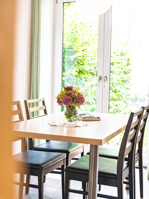 Ferienwohnung 6: Esstisch mit Blumenstrauß und Stühlen vor einer Terrassentür mit Blick ins Grüne