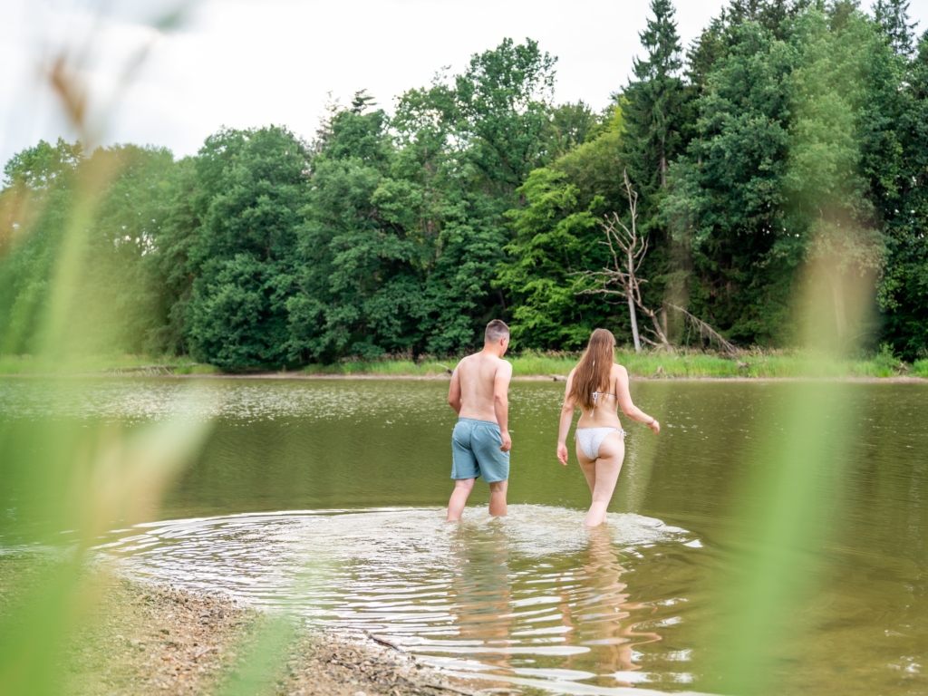 Zwei Erwachsene waten im flachen Wasser am Ufer des Neuweihers, dahinter Bäume