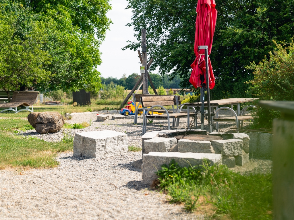 Grillplatz mit Granitblöcken und Sitzgelegenheiten im Grünen, im Hintergrund ein roter Sonnenschirm