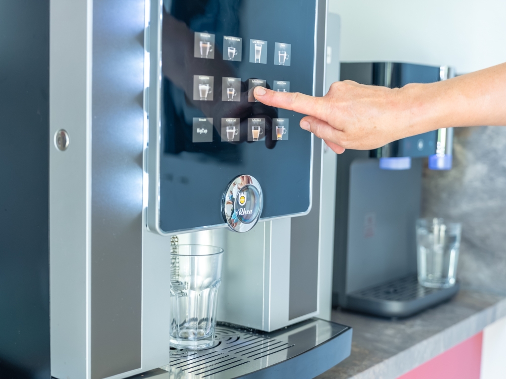 Hand drückt am Kaffeeautomaten im Hofladen eine Taste, darunter steht ein Glas auf der Ablage