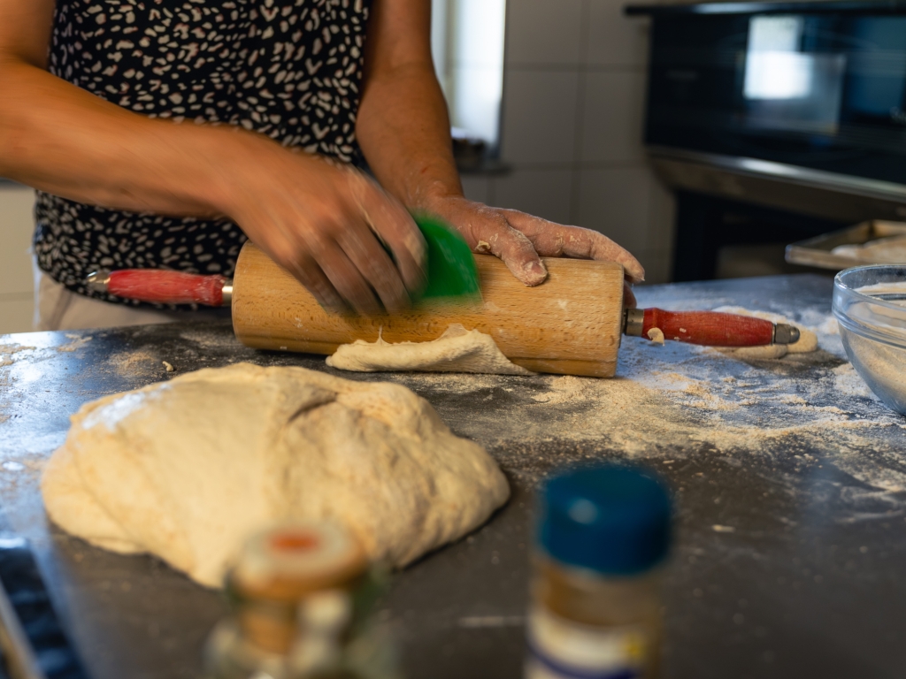 Beim Pizza backen: Teig wird mit dem Nudelholz ausgerollt, im Vordergrund ein Teigfladen