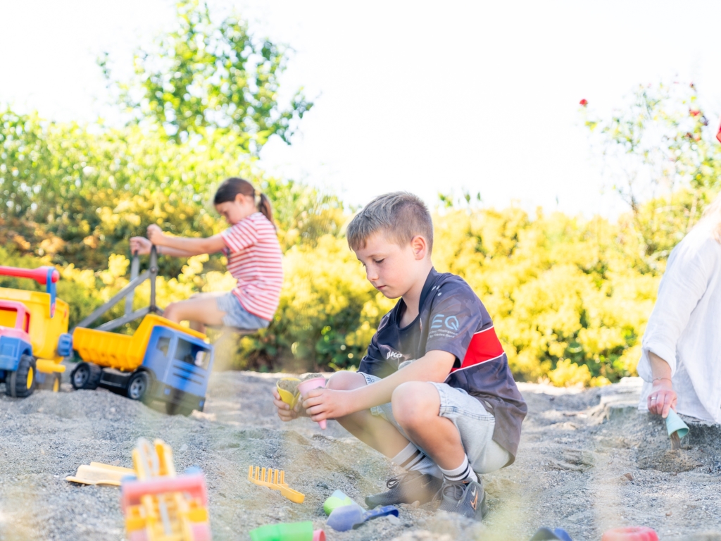 Zwei Kinder spielen im Sand mit Spielzeug, im Hintergrund gelb blühende Sträucher