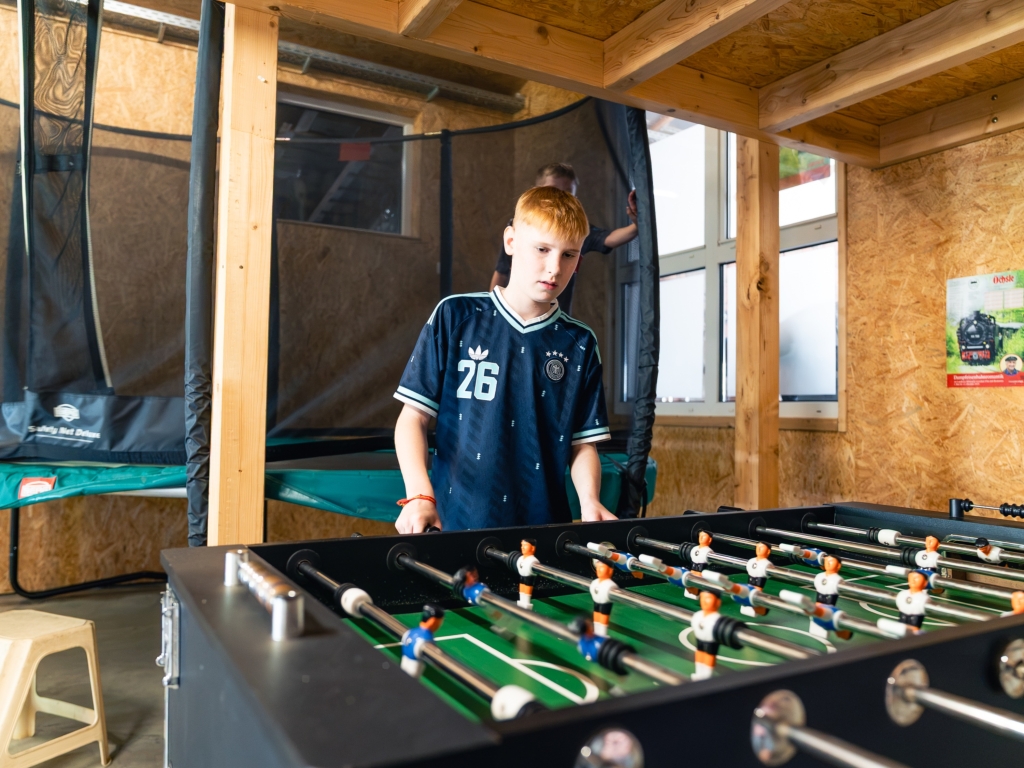 Zwei Jugendliche spielen in der Spielscheune am Kicker, im Hintergrund Trampolin