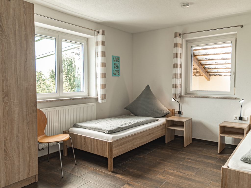 Ferienwohnung 10: Schlafzimmer mit Einzelbetten, hellem Holzbettrahmen, Stuhl und Fenstern mit Vorhängen