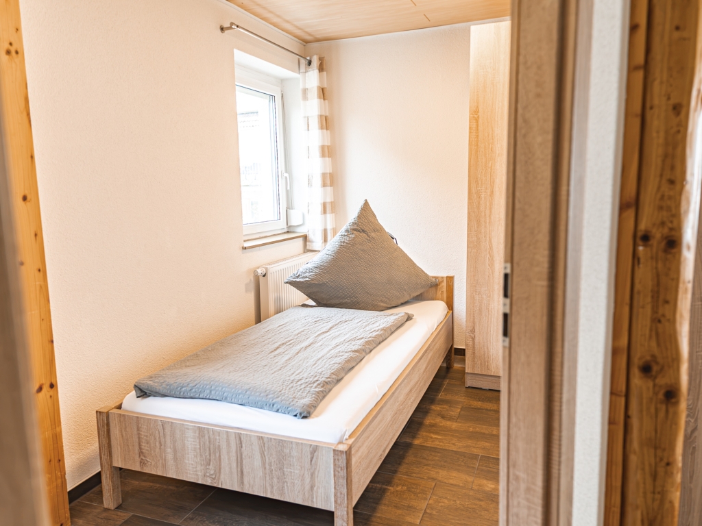 Ferienwohnung 10: Kleines Schlafzimmer mit Einzelbett, Kissen und Fenster, Blick durch die offene Tür