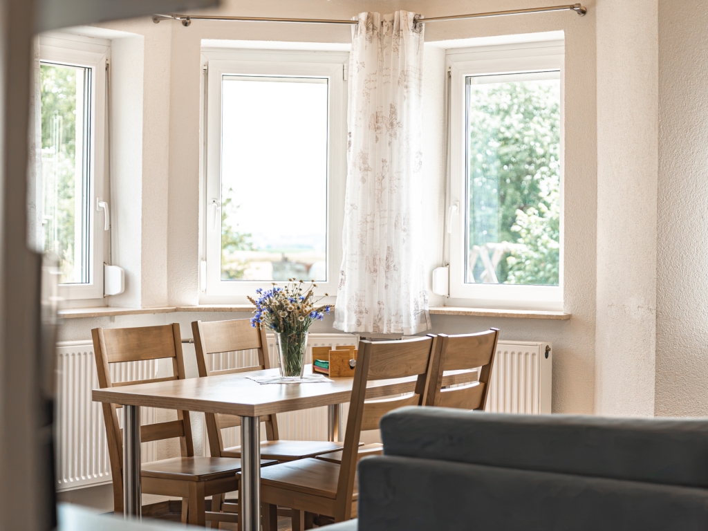 Ferienwohnung 10: Esstisch mit Holzstühlen vor dem Erkerfenster, Sofa im Vordergrund, Blick ins Grüne