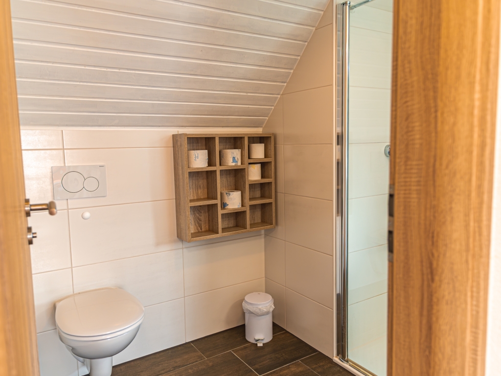 Ferienwohnung 11: WC-Raum mit Wand-WC, Holzregal an der Wand und Dachschräge mit heller Holzverkleidung