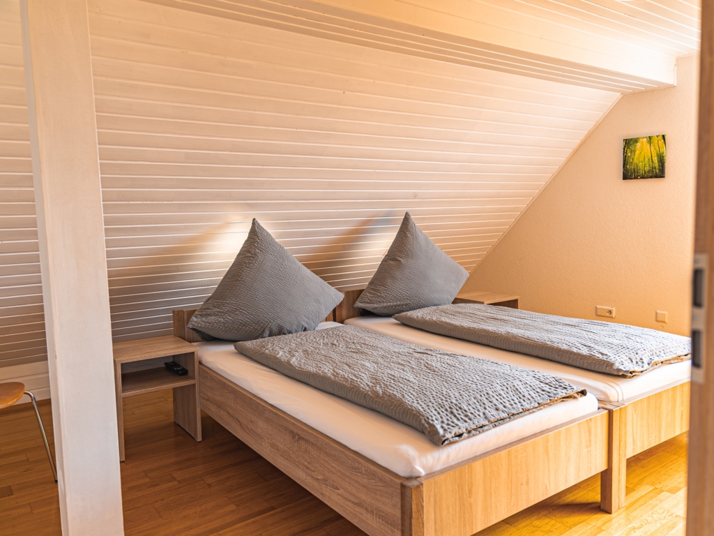 Ferienwohnung 11: Dachschlafzimmer mit zwei Betten, grauer Bettwäsche, Nachttisch und Holzverkleidung