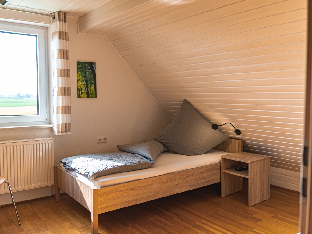 Ferienwohnung 11: Dachzimmer mit Einzelbett, Nachttisch, Fenster mit gestreiftem Vorhang und Holzverkleidung