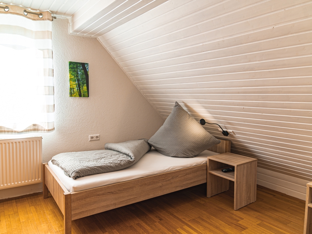 Ferienwohnung 11: Einzelbett mit grauem Kissen und Nachttisch unter der Dachschräge, Fenster mit Vorhang