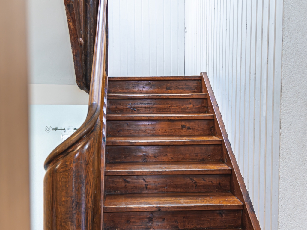 Ferienwohnung 11: Holztreppe mit dunklem Geländer und heller Wandverkleidung im Treppenhaus