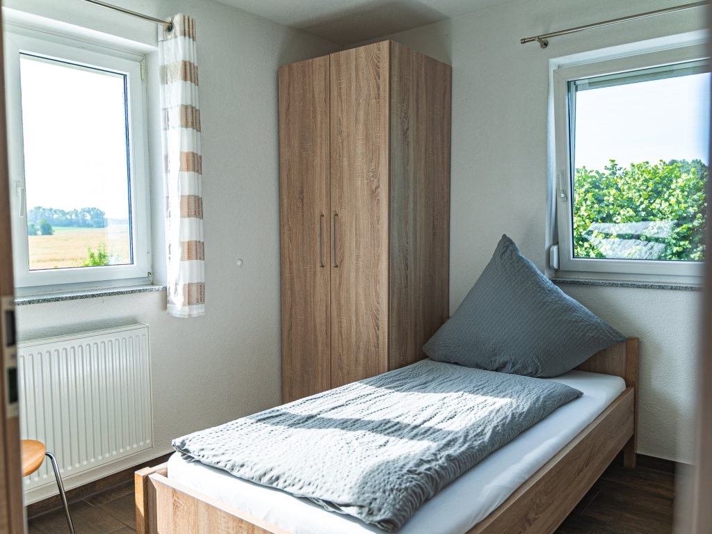 Ferienwohnung 11: Schlafzimmer mit Einzelbett, Holzkleiderschrank und zwei Fenstern, Blick ins Grüne