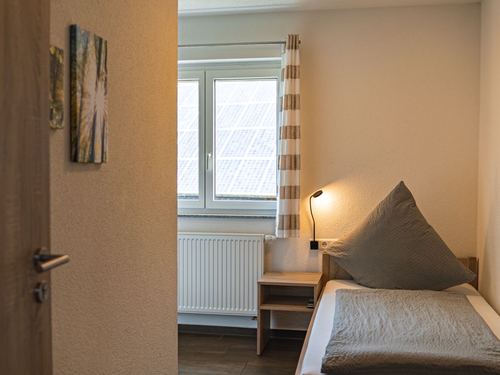 Ferienwohnung 11: Schlafzimmer mit Einzelbett, Nachttisch mit Lampe und Fenster mit gestreiftem Vorhang