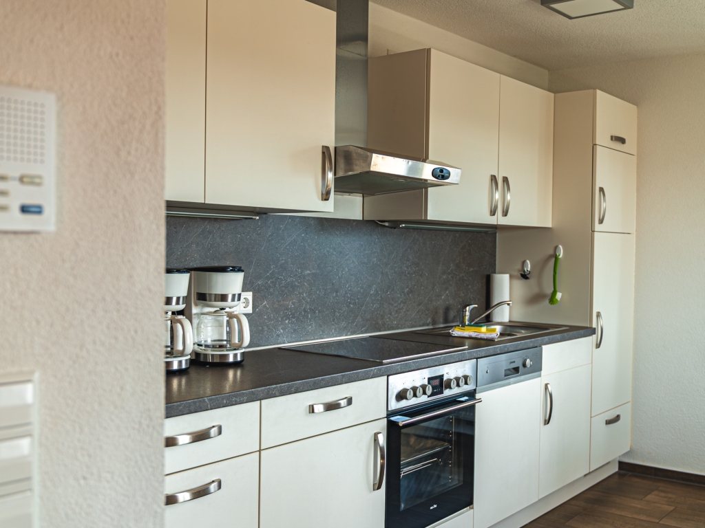 Ferienwohnung 11: Küchenzeile mit weißen Schränken, Herd, Backofen, Dunstabzug und dunklem Spritzschutz
