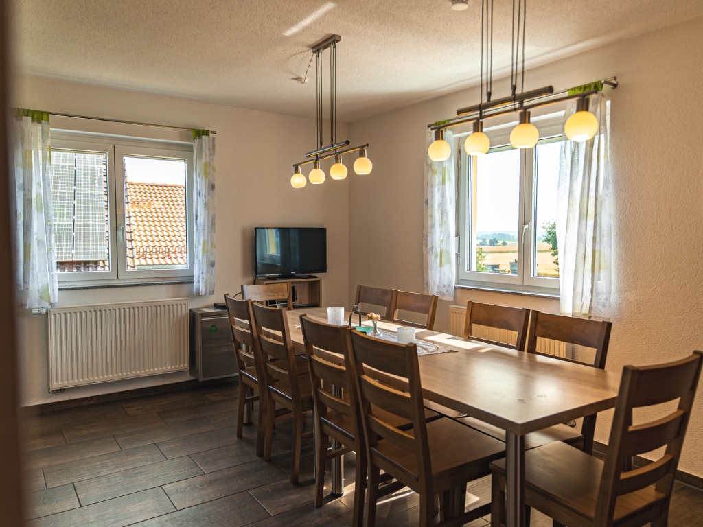 Ferienwohnung 11: Langer Esstisch mit Holzstühlen, Hängelampen, Fenstern mit Vorhängen und Fernseher