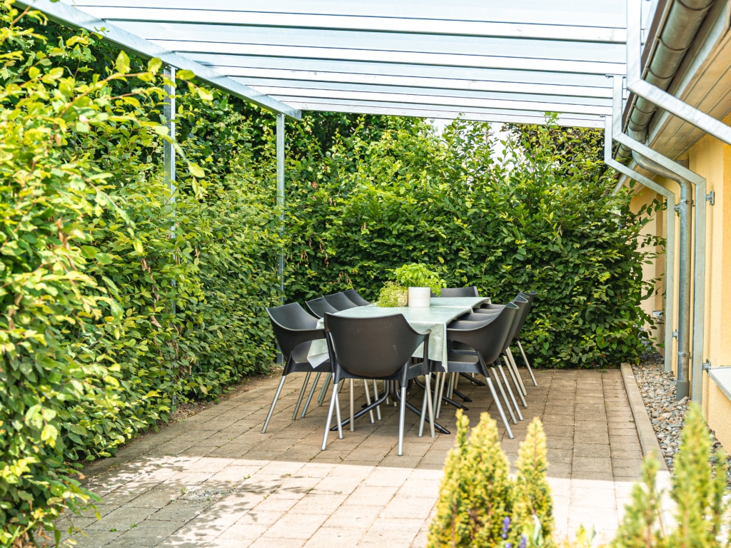 Ferienwohnung 11: Überdachte Terrasse mit Tisch und dunklen Stühlen, umgeben von Hecken