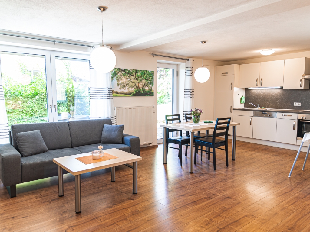 Ferienwohnung 1: Wohnbereich mit grauem Sofa, Esstisch und offener Küche vor bodentiefen Fenstern
