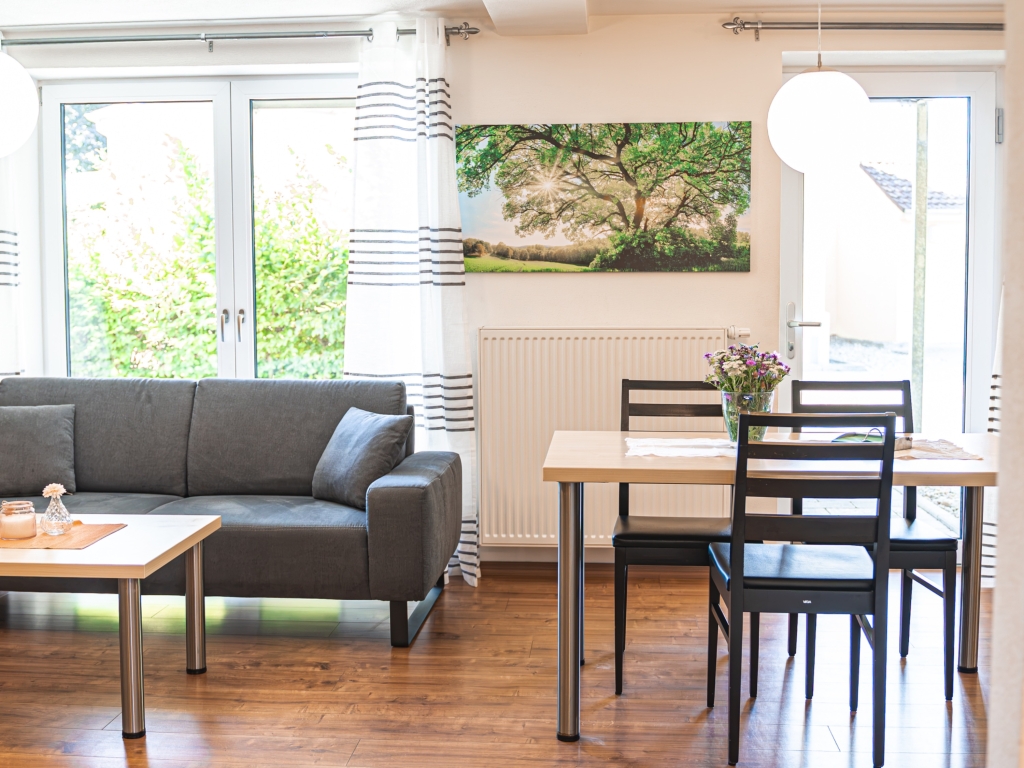 Ferienwohnung 1: Wohn-Essbereich mit Sofa, Esstisch und Baumbild, bodentiefe Fenster zum Grünen