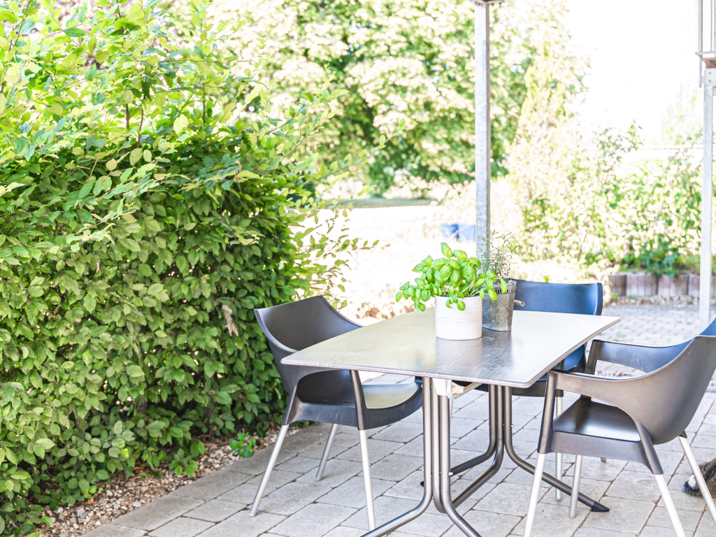 Ferienwohnung 1: Terrasse mit Tisch und Stühlen, daneben eine dichte grüne Hecke und Kräuter im Topf