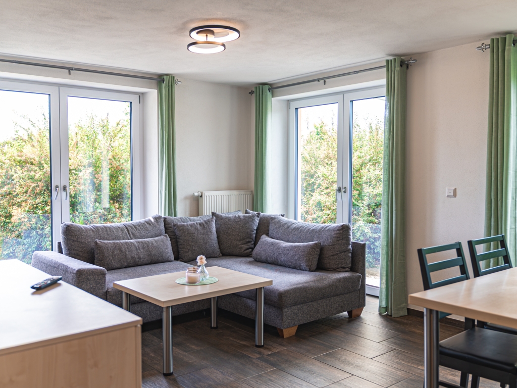 Ferienwohnung 2: Wohnbereich mit grauem Sofa, Couchtisch und zwei bodentiefen Fenstertüren ins Grüne