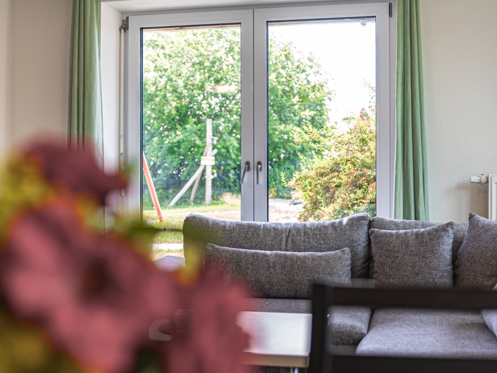 Ferienwohnung 2: Blick vom Wohnbereich durch das Fenster in den Garten, im Vordergrund Blumen und Sofa