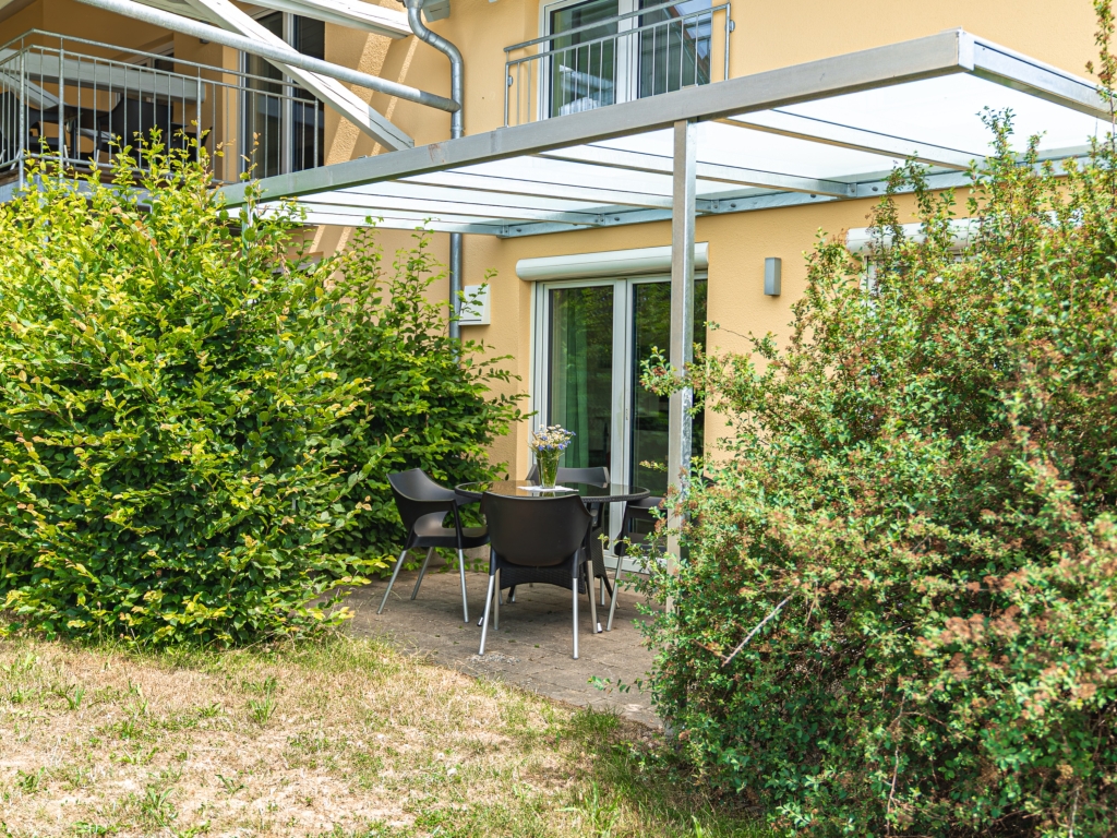 Ferienwohnung 2: Überdachte Terrasse mit Tisch und Stühlen, umgeben von Hecken und Rasen