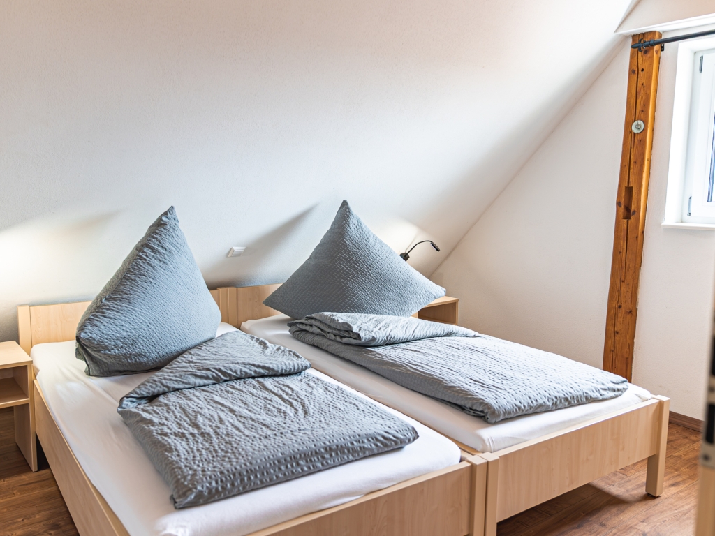 Ferienwohnung 3: Schlafzimmer unter der Dachschräge mit zwei Betten, grauer Bettwäsche und Leselampe