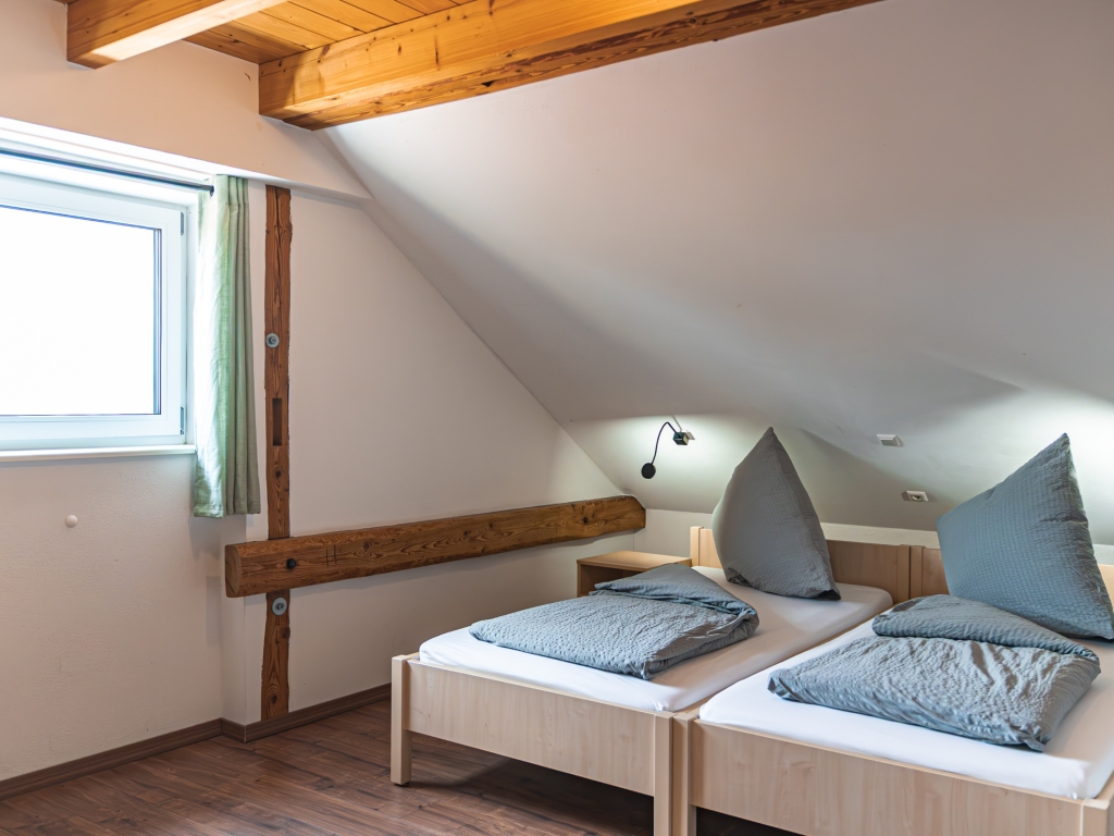 Ferienwohnung 3: Dachgeschoss-Schlafzimmer mit zwei Betten, Holzbalken, Fenster und grünem Vorhang