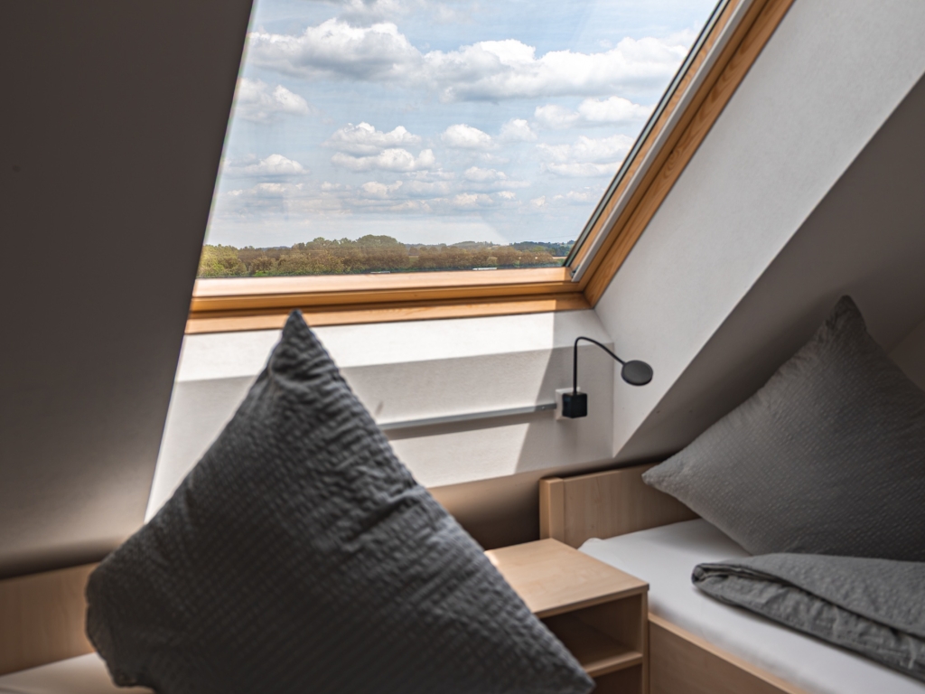 Ferienwohnung 3: Blick aus dem Dachfenster des Schlafzimmers über Felder und Himmel, davor Kissen und Bett