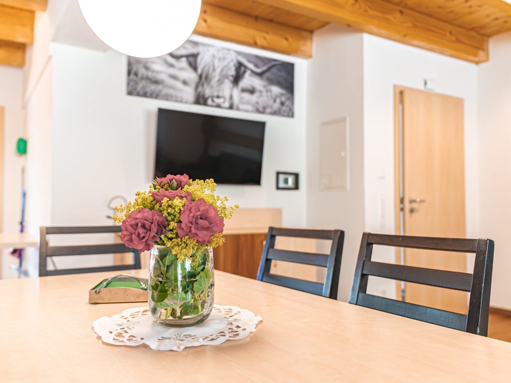 Ferienwohnung 3: Esstisch mit Blumenstrauß, dahinter Fernseher an der Wand und Holztür