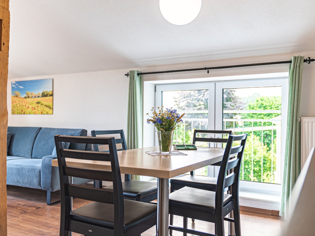 Ferienwohnung 4: Esstisch mit Stühlen und Blumenstrauß, daneben blaues Sofa und Fenster zum Balkon