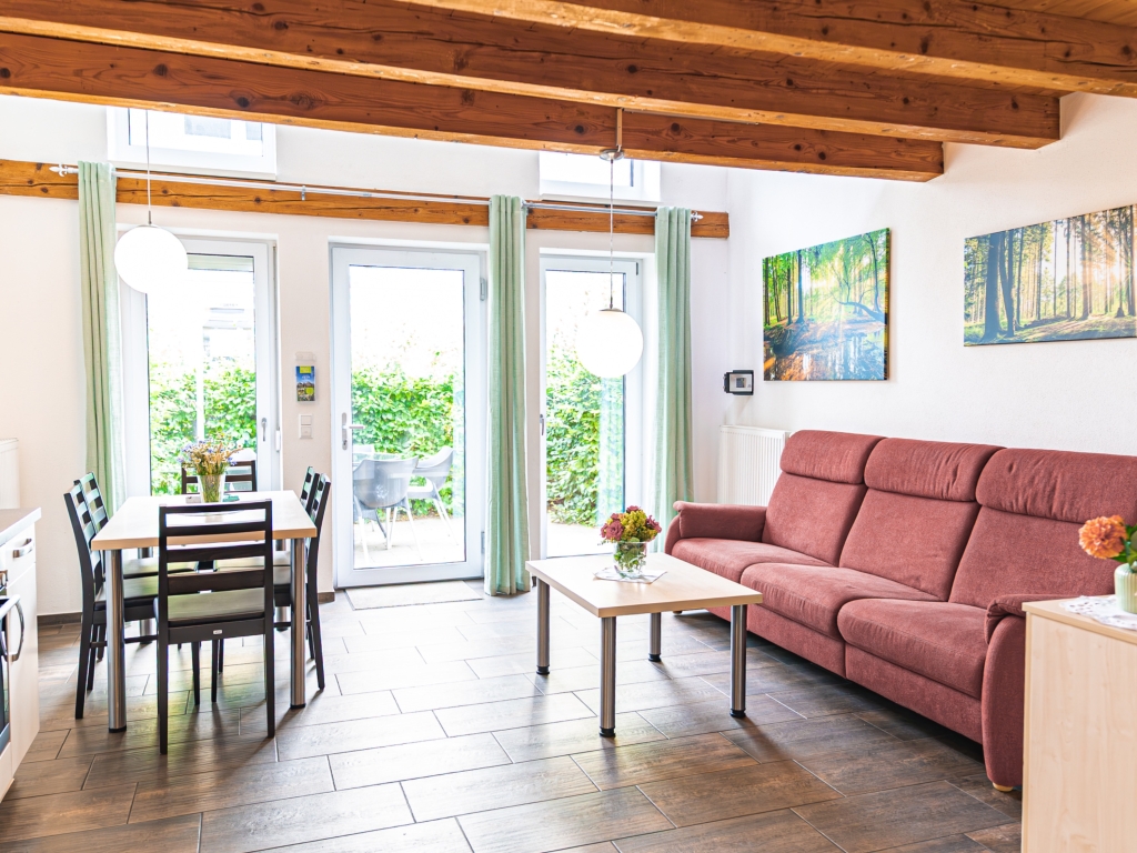 Ferienwohnung 5: Wohn-Essbereich mit rotem Sofa, Couchtisch, Esstisch und Terrassentüren mit Blick ins Grüne