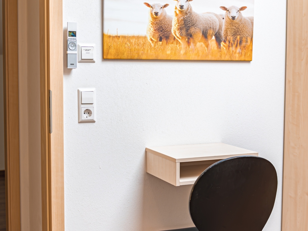 Ferienwohnung 6: Kleine Ablage an der Wand mit Stuhl, darüber ein Wandbild mit Schafen auf einer Wiese