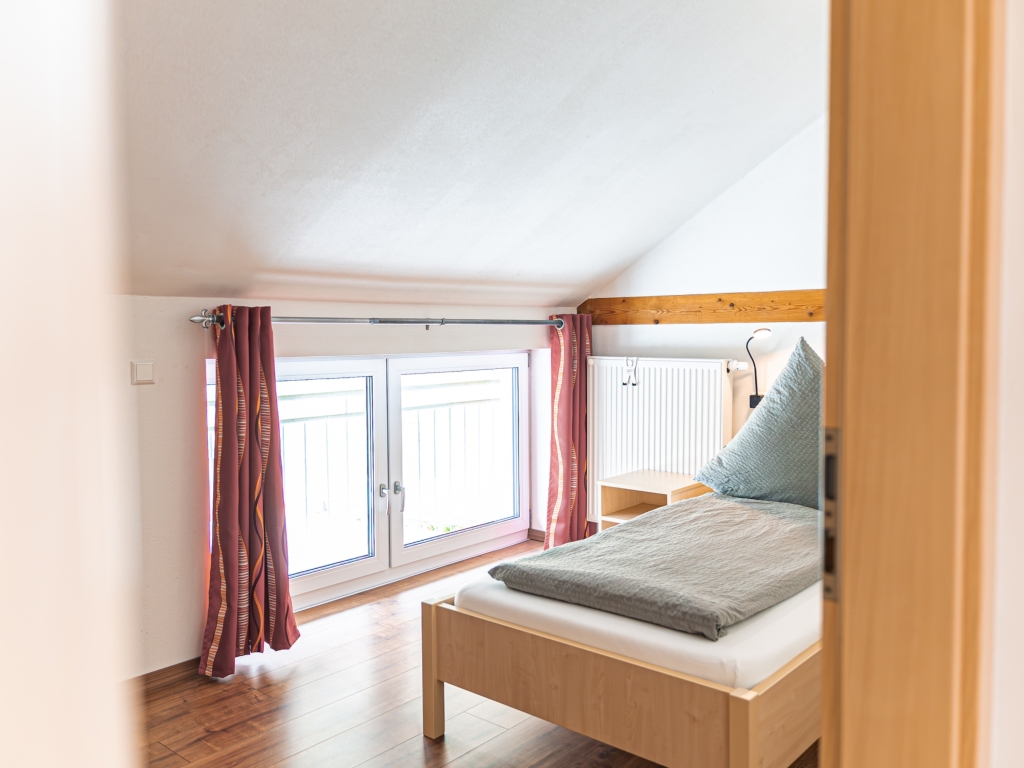 Ferienwohnung 6: Schlafbereich mit Einzelbett unter der Dachschräge, daneben Fenster mit rotem Vorhang
