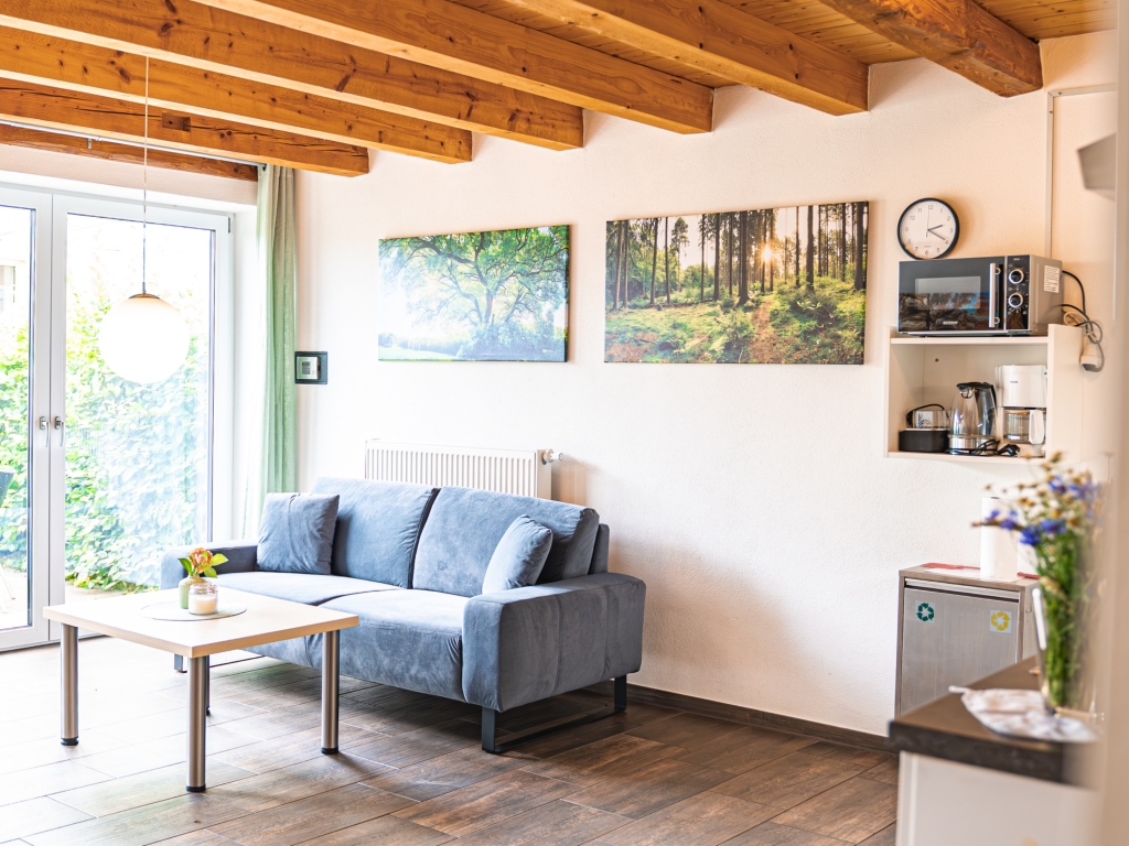 Ferienwohnung 6: Wohnbereich mit blauem Sofa, Couchtisch, Holzbalkendecke und Waldbild an der Wand