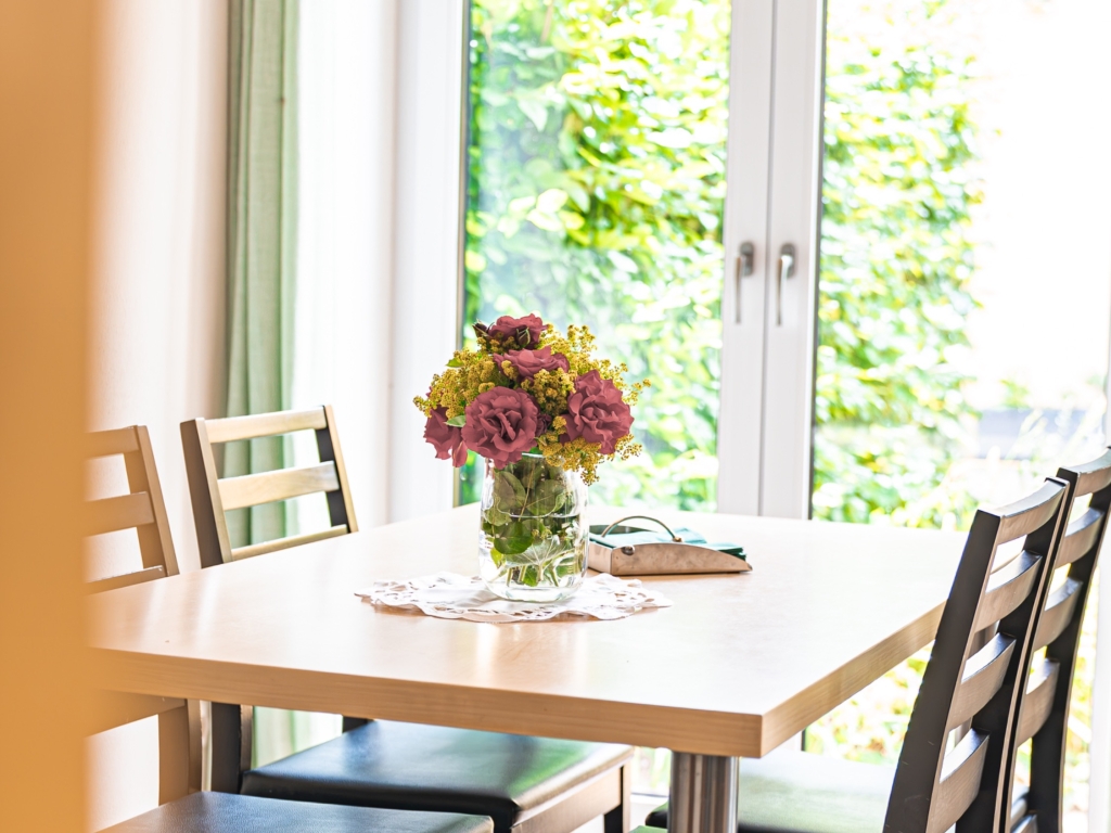 Ferienwohnung 6: Esstisch mit Blumenstrauß und Stühlen vor einer Terrassentür mit Blick ins Grüne