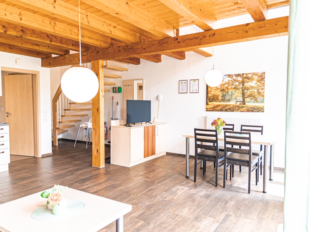 Ferienwohnung 6: Offener Wohn-Essbereich mit Esstisch, Fernseher und Holztreppe zum Obergeschoss