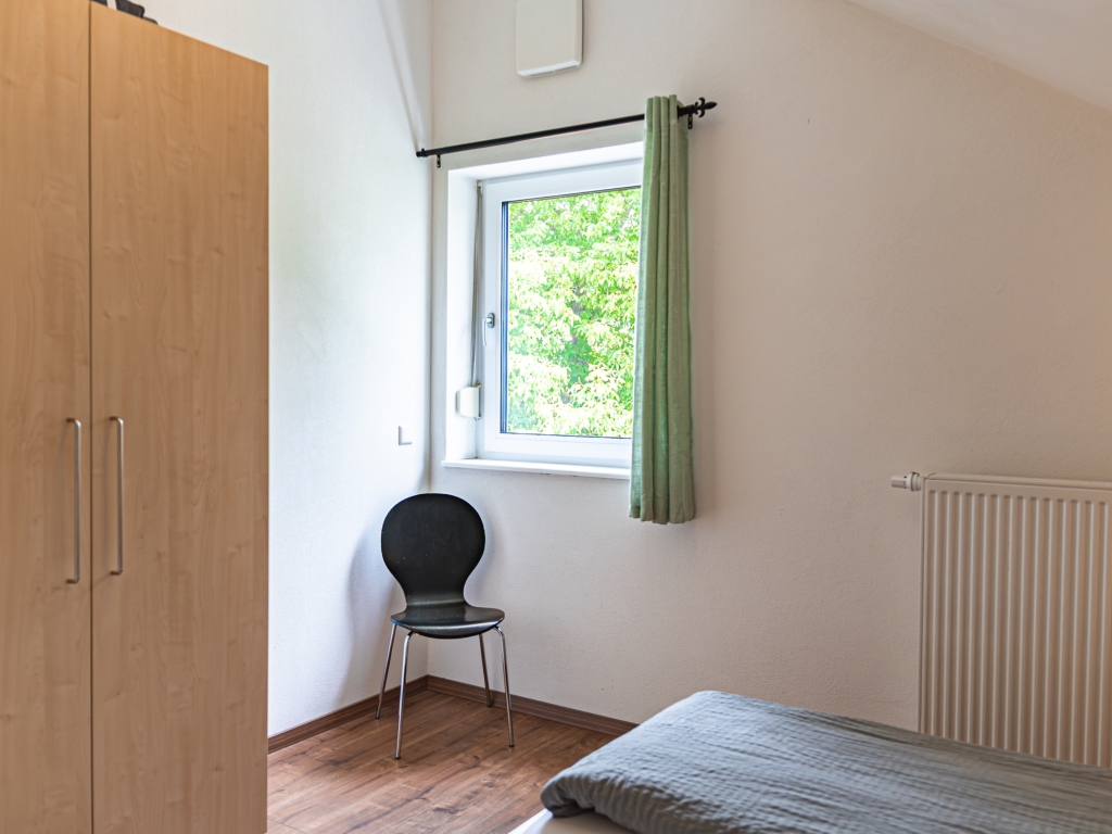 Ferienwohnung 7: Schlafzimmer mit Kleiderschrank, Stuhl und Fenster mit grünem Vorhang unter der Dachschräge
