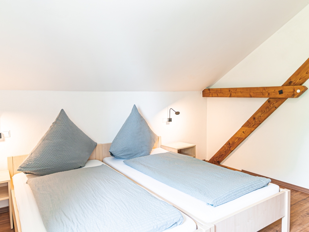 Ferienwohnung 7: Schlafzimmer mit zwei Betten, Nachttisch mit Leselampe und sichtbarem Dachbalken